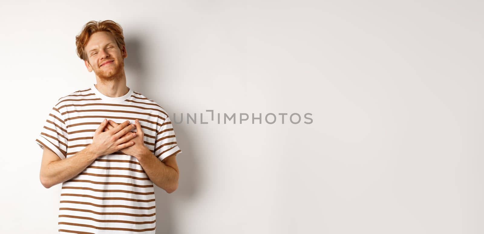 Happy young man with ginger hair appreciate gift, holding hands on heart and smiling, thanking you, standing grateful against white background.