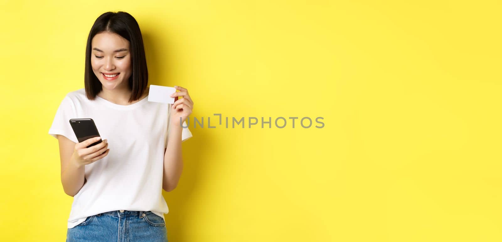E-commerce and online shopping concept. Beautiful asian woman paying in internet, looking at smartphone screen and holding plastic credit card, yellow background by Benzoix