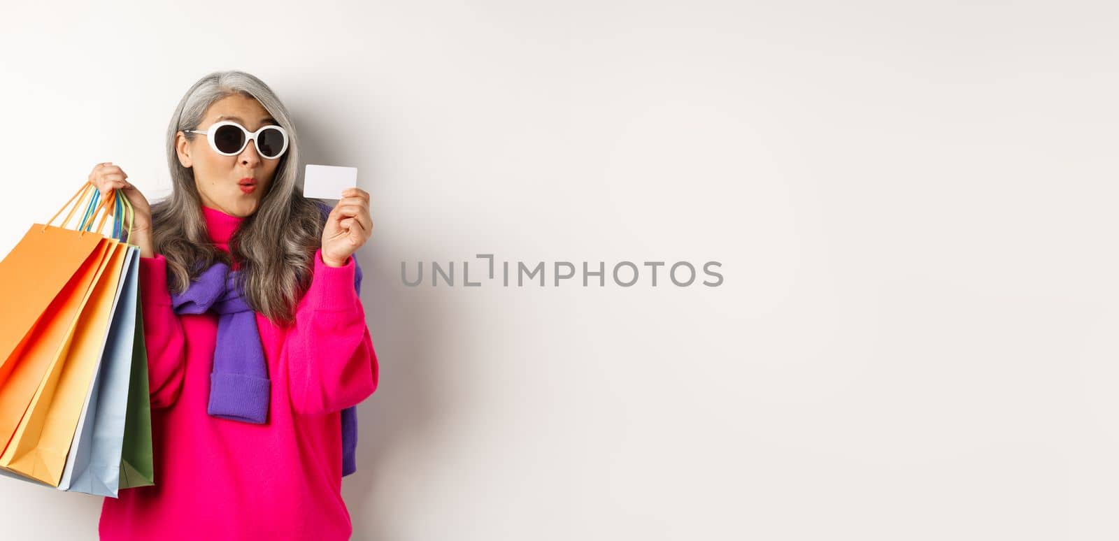 Fashionable asian old lady going shopping, holding paper bags and plastic credit card, wearing stylish sunglasses, standing over white background.