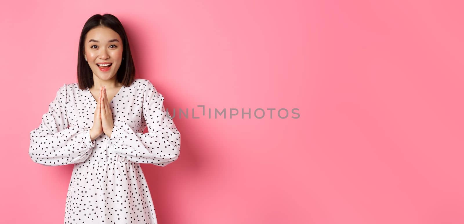 Beautiful asian woman thanking you, holding hands together in appreciation gesture, smiling happy at camera, standing grateful over pink background.