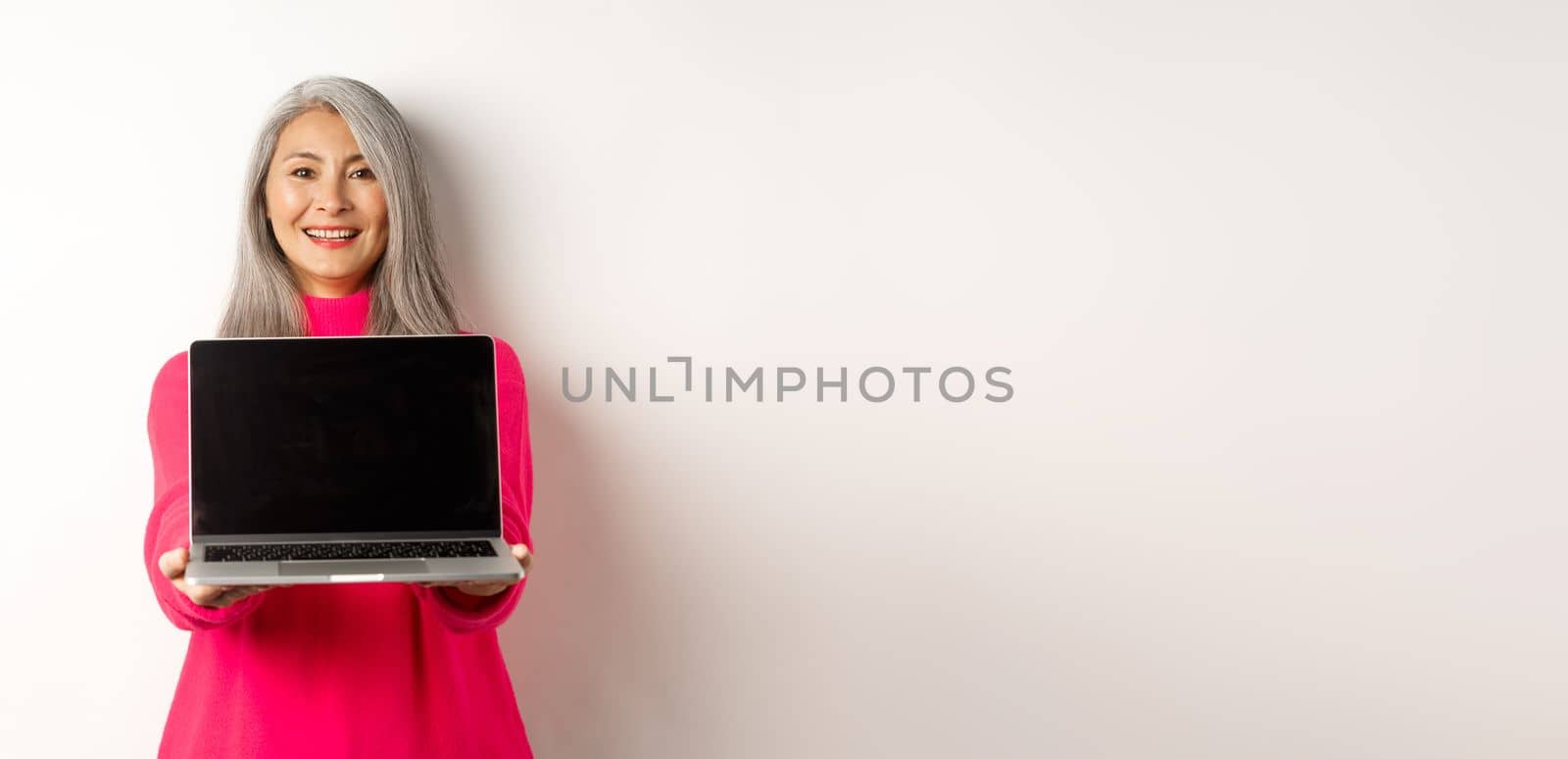 E-commerce concept. Smiling asian senior woman showing blank laptop screen and looking happy, demonstrating promo, white background.