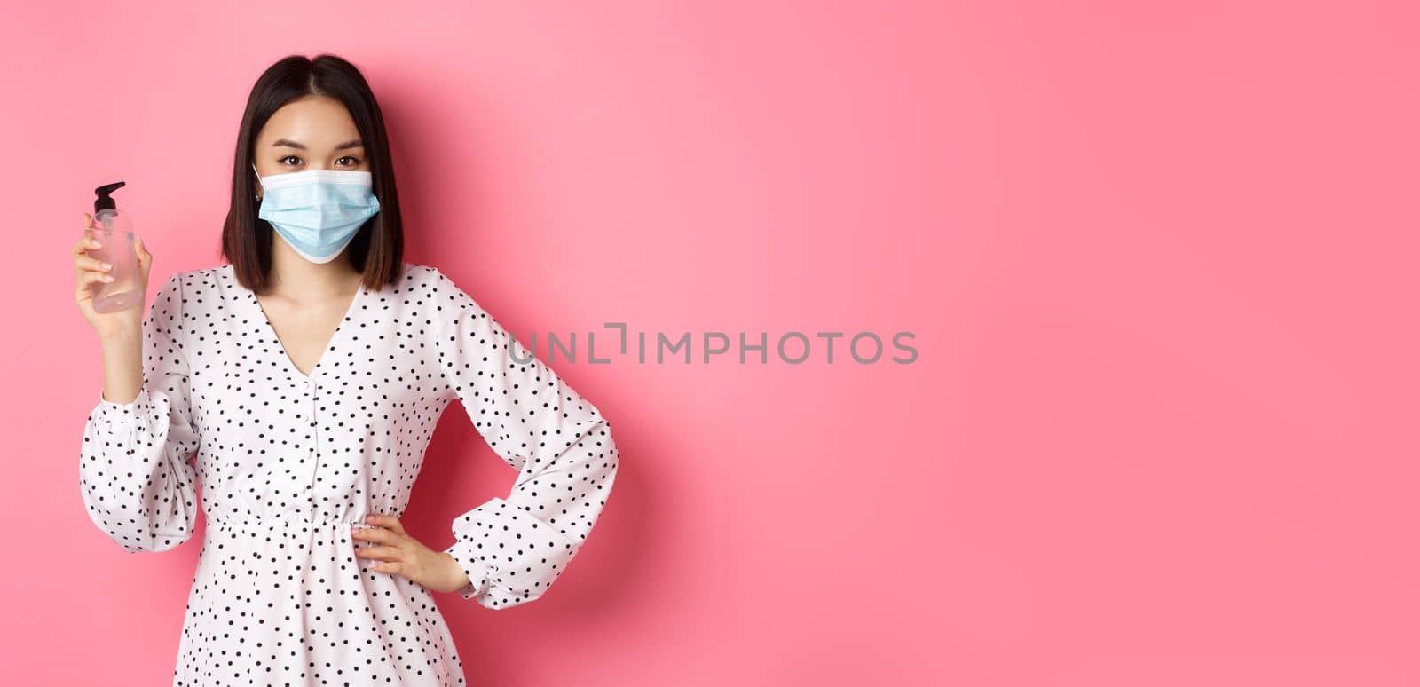 Covid-19, pandemic and lifestyle concept. Beautiful korean woman in dress and medical mask showing hand sanitizer, recommending antiseptic, standing over pink background.