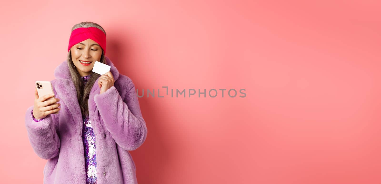 Online shopping and fashion concept. Stylish asian grandmother making purchase in internet with smartphone and plastic credit card, pink background.