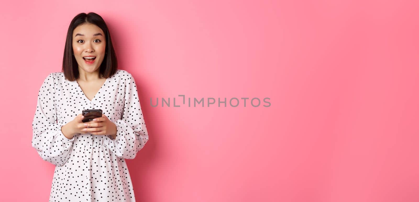 Online shopping. Amazed asian woman looking at camera with happy smile, making purchase with smartphone, using mobile phone app, standing over pink background.