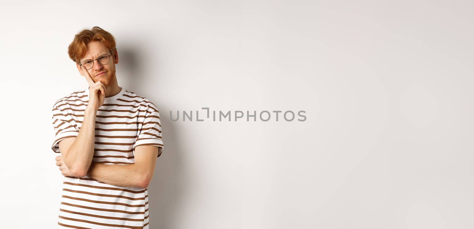 Young man with red messy hair and glasses looking doubtful, thinking and squinting at camera, standing thoughtful over white background.