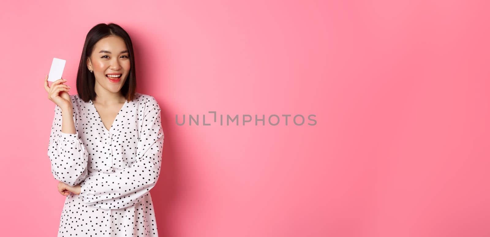 Shopping concept. Confident and happy asian woman holding credit card and smiling satisfied, standing against pink background by Benzoix