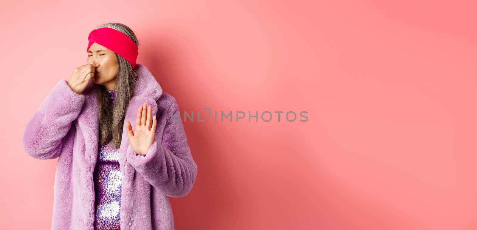 Disgusted asian mature woman in trendy purple winter coat and dress, shut nose and showing stop, refusal gesture, smell something disgusting, standing over pink background.