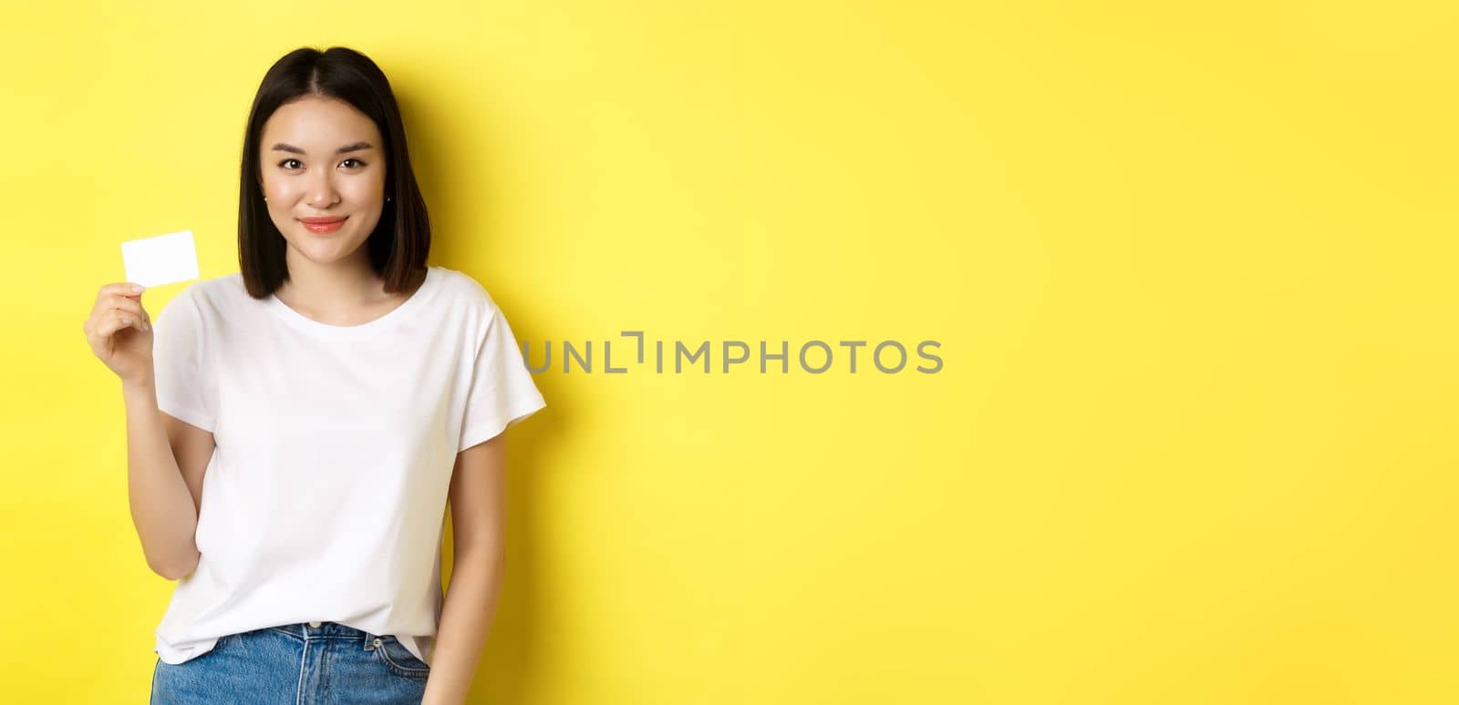 Young asian woman in casual white t-shirt showing plastic credit card and smiling at camera, yellow background by Benzoix