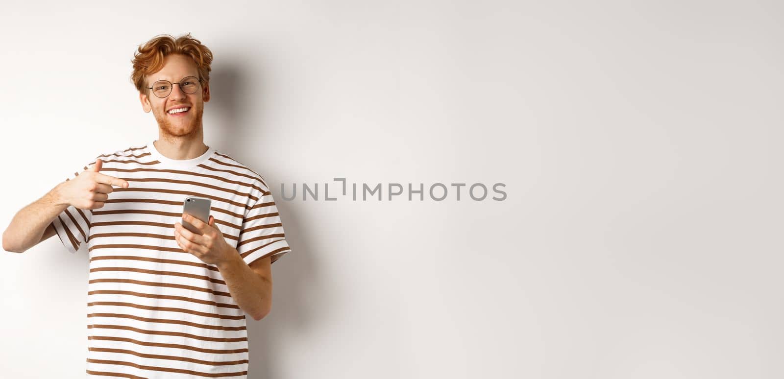 Technology and e-commerce concept. Young man with red messy hair, wearing glasses and t-shirt, pointing finger at smartphone and smiling satisfied, white background by Benzoix