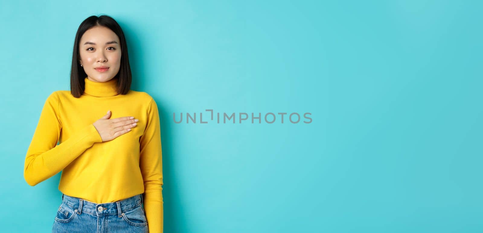 Image of proud smiling asian woman holding hand on heart, showing respect to national anthem, standing over blue background by Benzoix