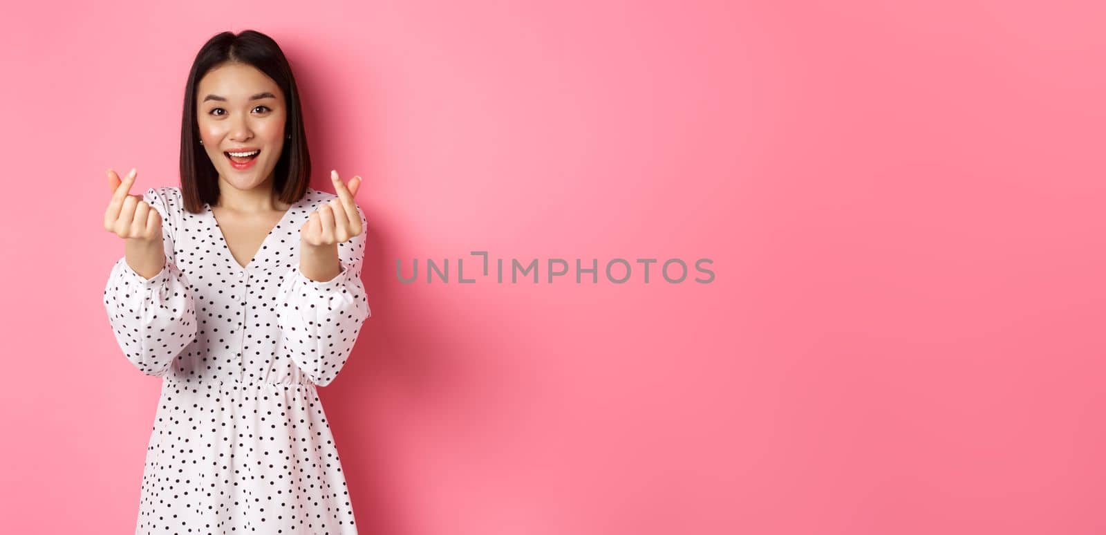 Lovely asian woman in dress showing korean heart signs and smiling, standing on romantic pink background.