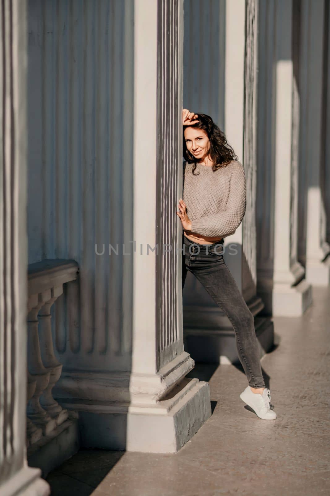 Woman building columns. An athletic woman in her 40s, dressed in a beige sweater and black jeans, poses near the pillars of a building. Walking around the city, tourism
