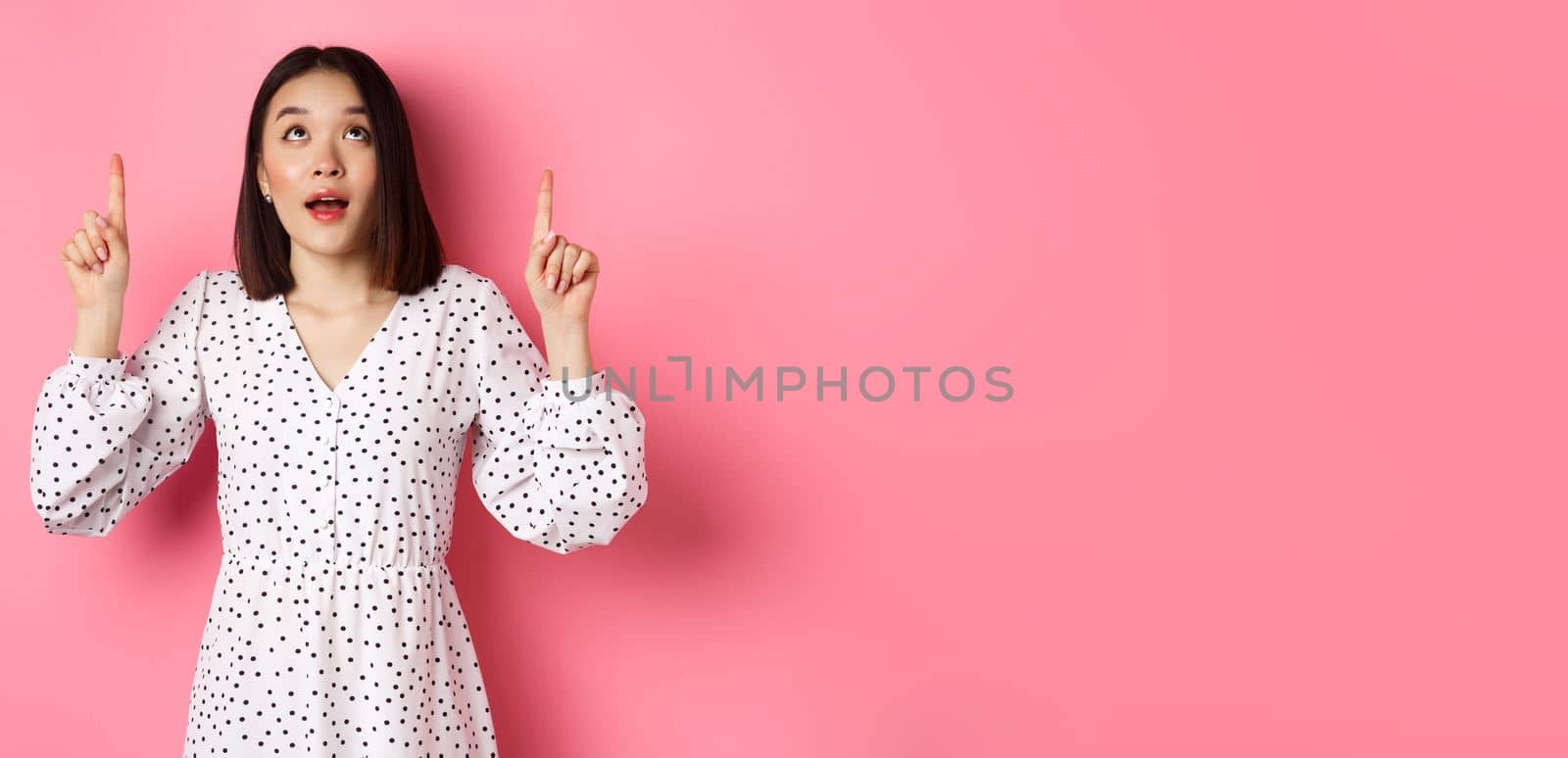 Image of beautiful asian girl in dress looking, pointing fingers up, gazing dreamy at promo offer, standing over pink background.