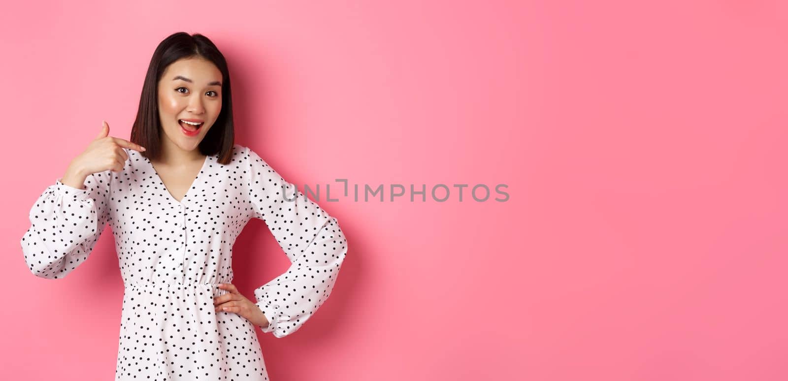 Beautiful asian girl in dress looking happy, pointing finger at herself, standing on romantic pink background by Benzoix