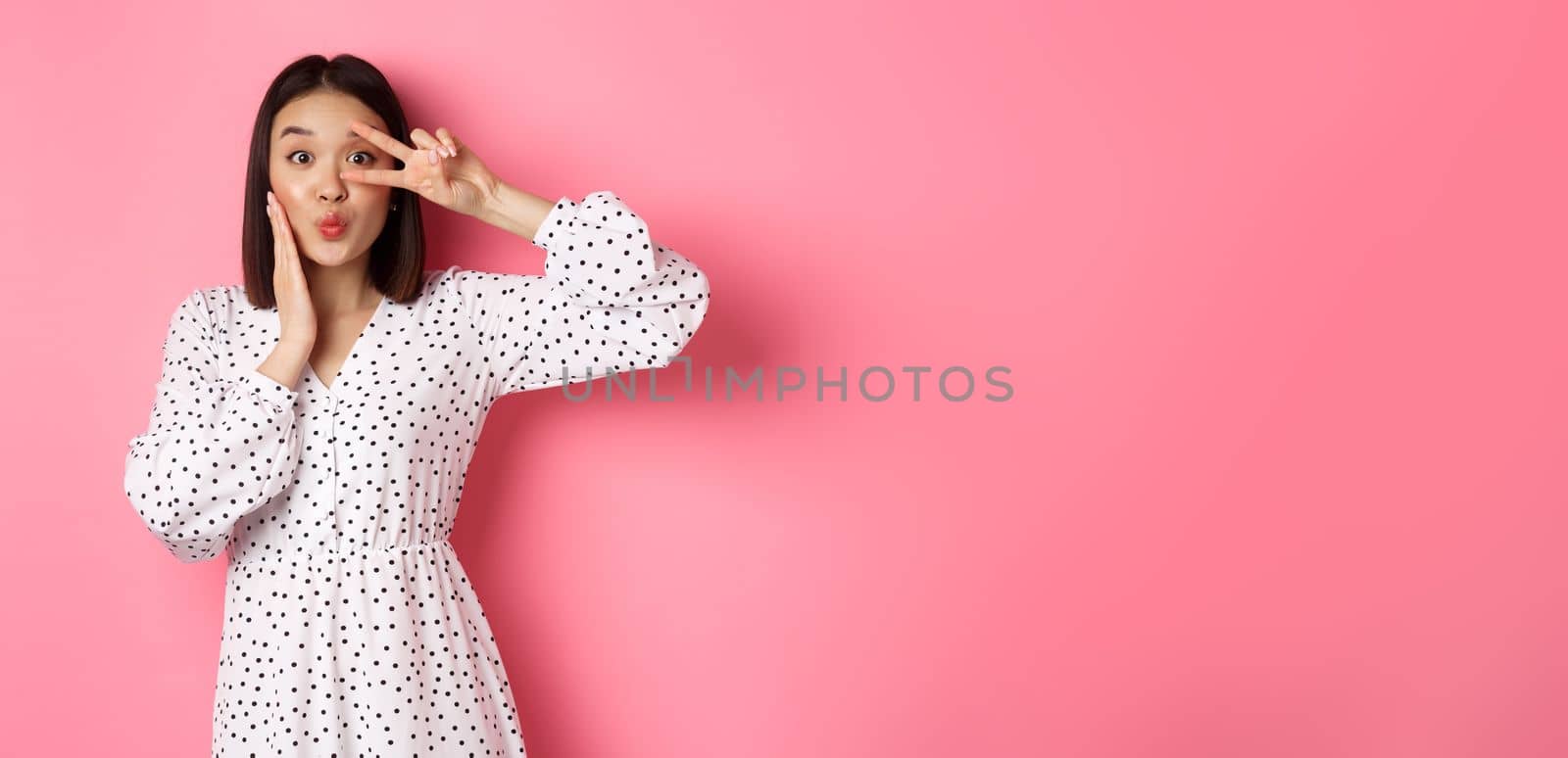 Coquettish asian woman in dress showing peace sign on eye, pucker lips for kiss, standing silly against pink background by Benzoix