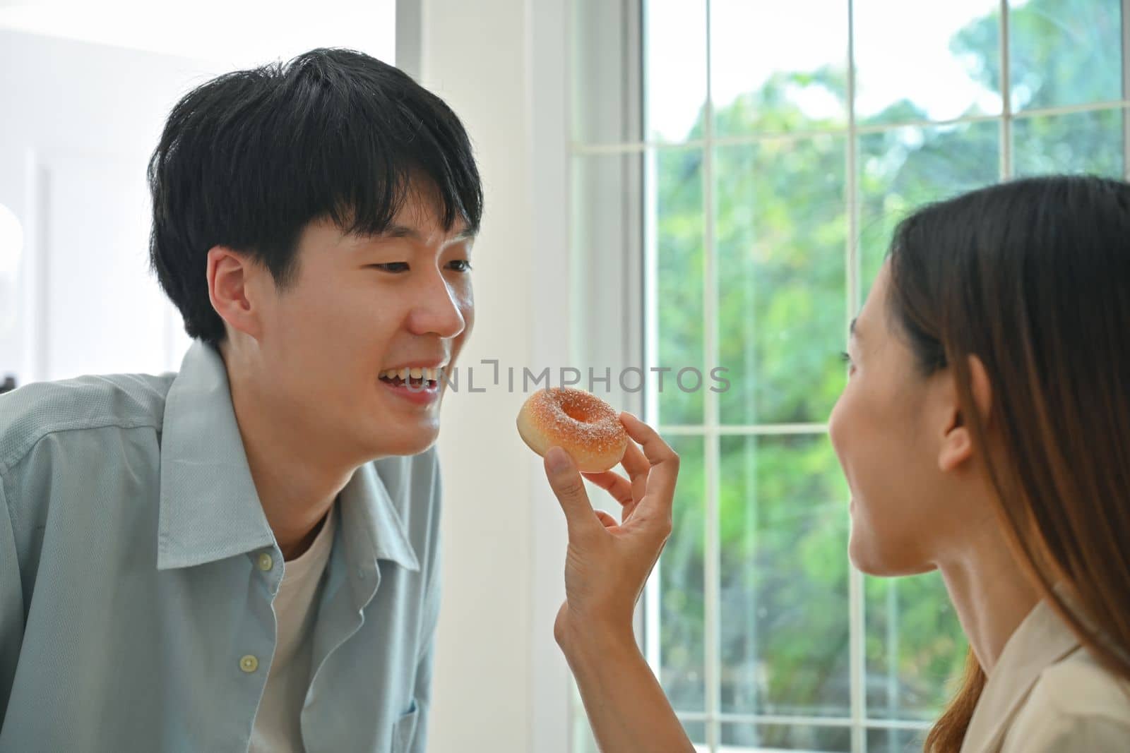 Young woman feeding his boyfriends with donut while cooking in modern kitchen at home, enjoying leisure weekend time together by prathanchorruangsak