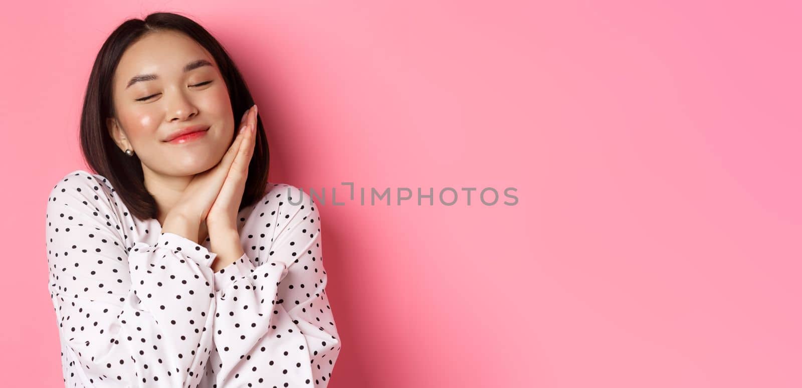 Beauty and lifestyle concept. Close-up of beautiful and dreamy asian woman sleeping on her hands, close eyes and smiling, daydreaming on pink background.