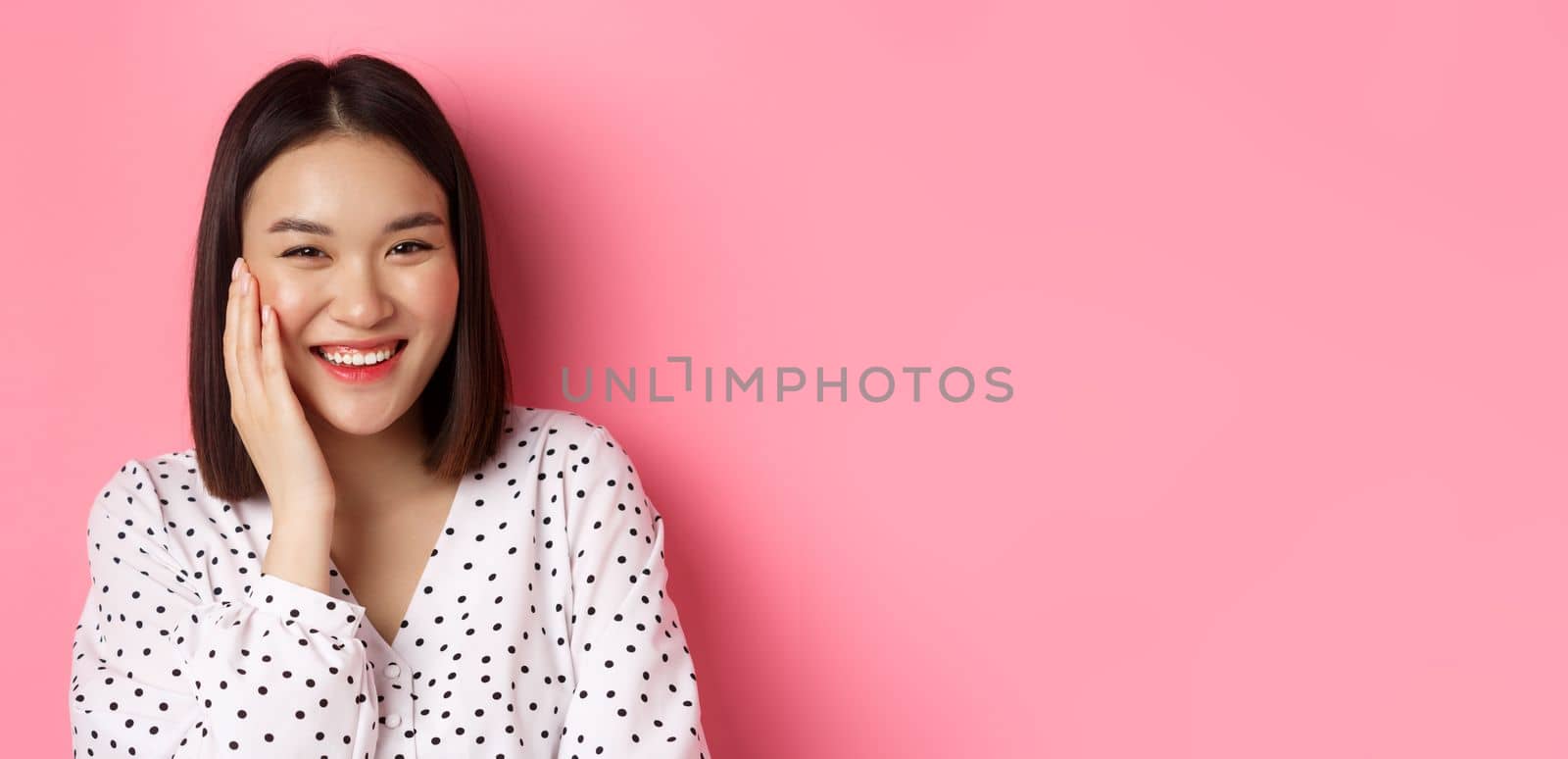 Close-up of adorable asian woman blushing, touching cheek and smiling cute at camera, standing over pink background.