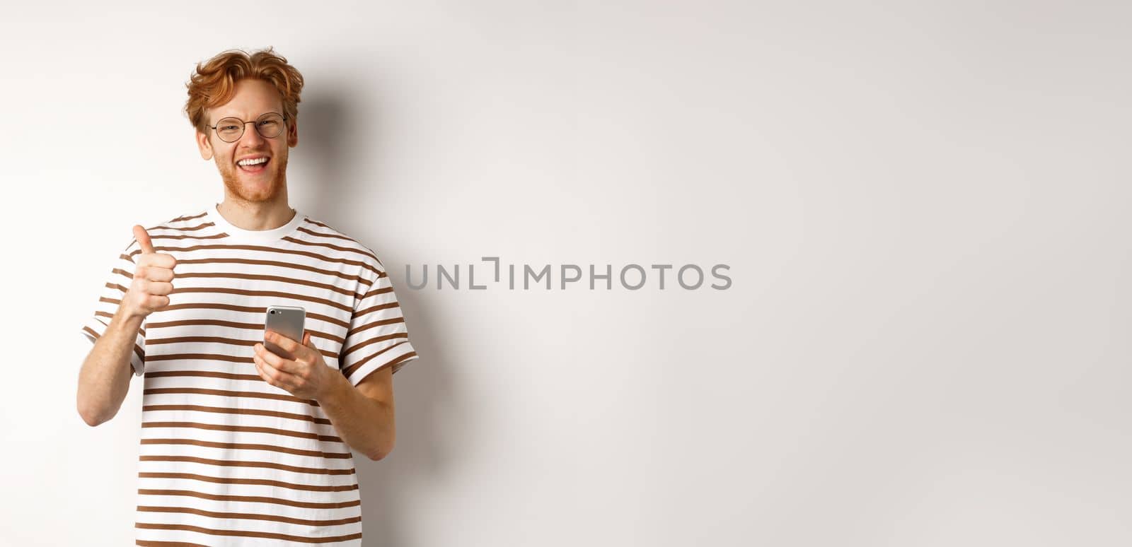 Technology and e-commerce concept. Satisfied male model with red hair, showing thumbs-up and holding smartphone, white background.
