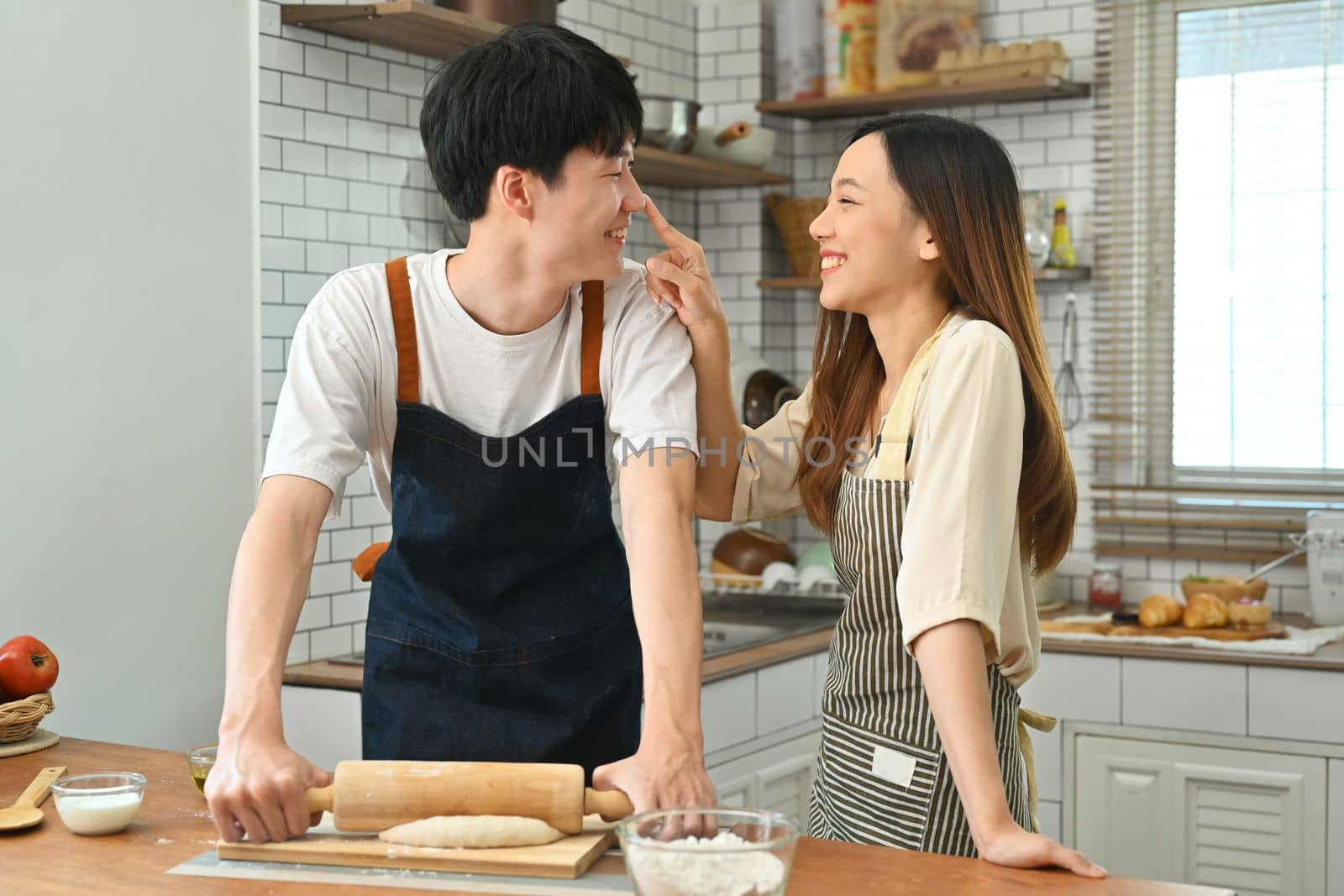 Smiling Asian man kneading dough with a rolling pin on wooden table, talking with girlfriend in kitchen by prathanchorruangsak