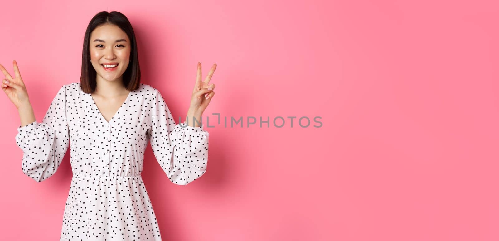 Cute asian brunette girl in dress smiling, showing kawaii peace signs and looking happy, standing over pink background by Benzoix