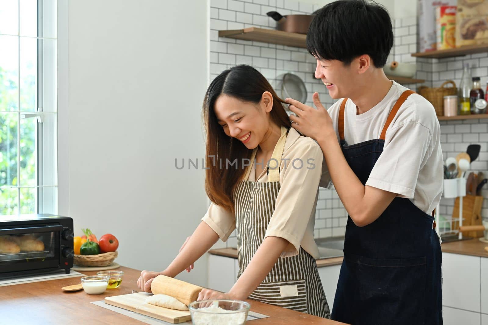 Romantic young couple preparing homemade pastry, kneading dough with a rolling pin on wooden table in kitchen.