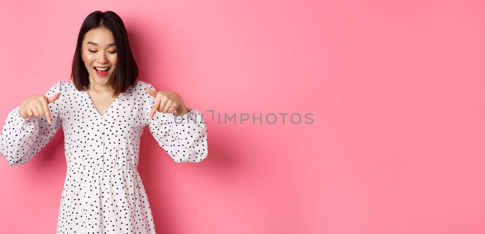 Romantic asian woman peeking down, pointing fingers at bottom, looking curious at product discount with happy smile, standing over pink background.