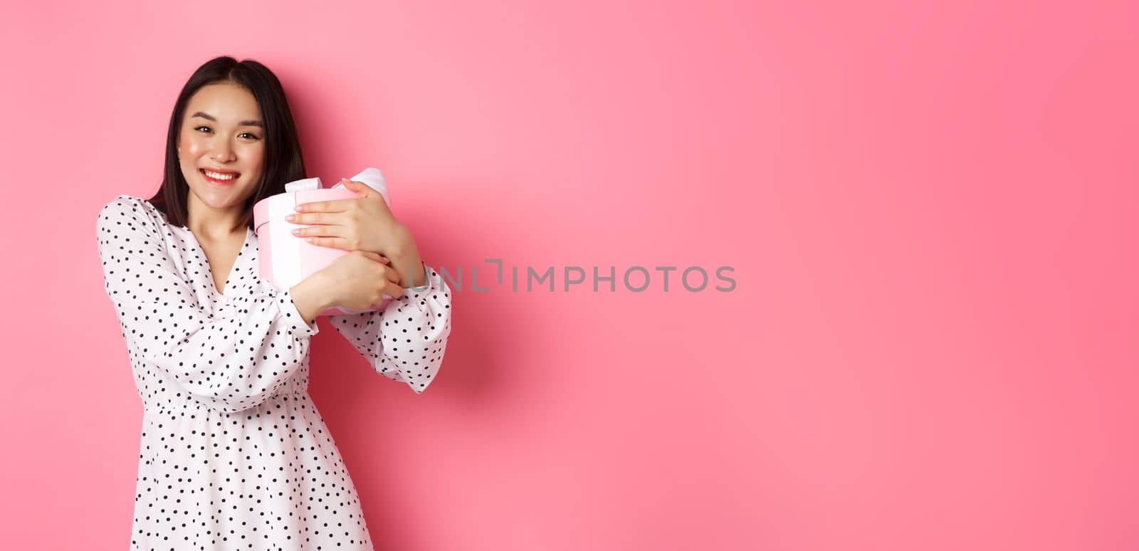 Lovely asian woman hugging her gift and smiling thankful, receive valentines day present, standing over pink background by Benzoix