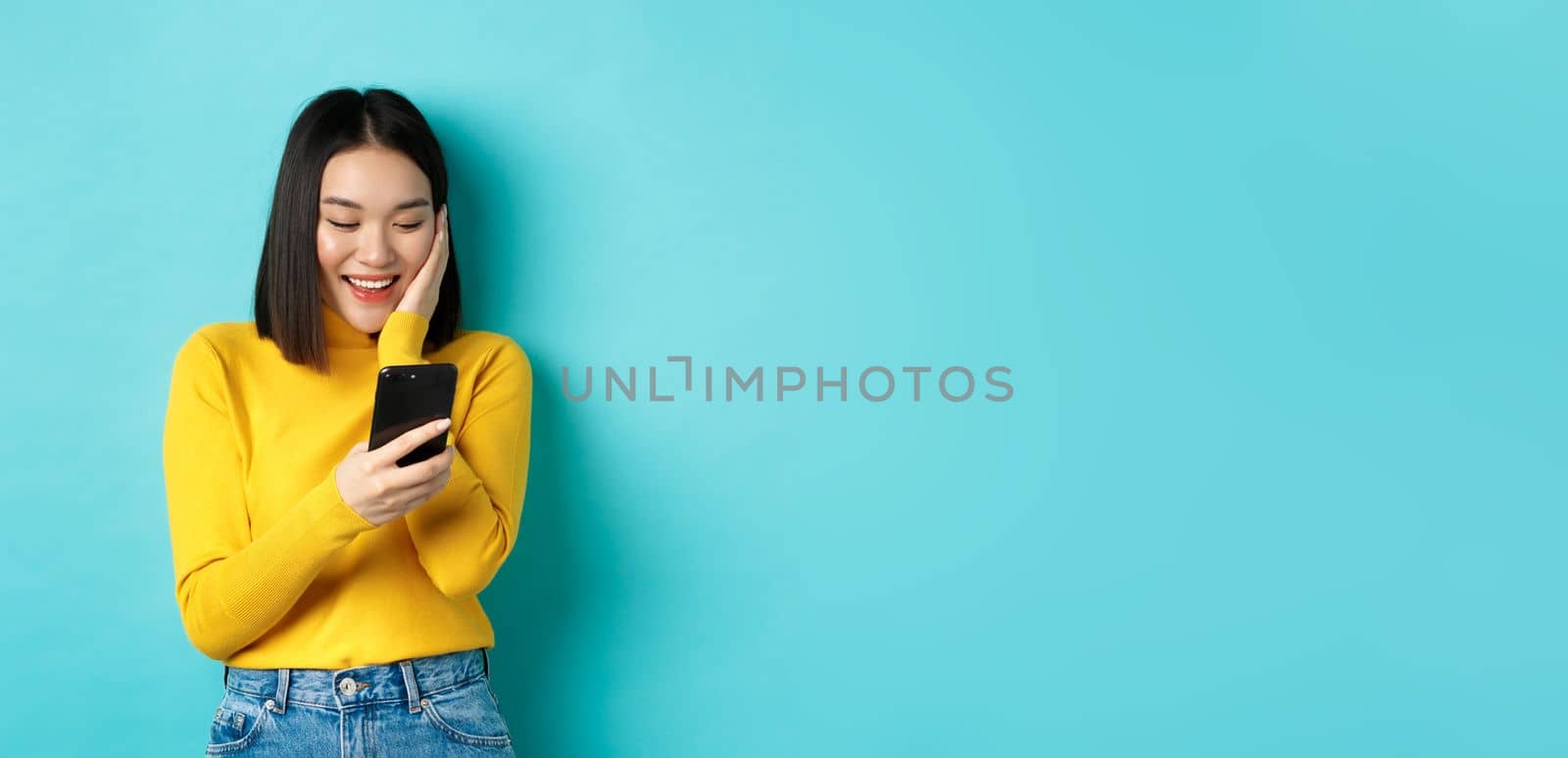 Image of happy asian woman reading message on mobile phone screen and smiling, chat in smartphone app, standing over blue background.