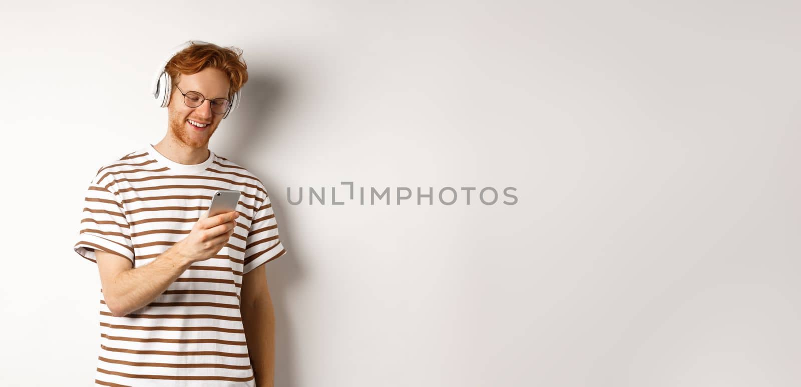 Technology concept. Young redhead hipster in glasses, listening music in headphoes, reading message on smartphone, white background by Benzoix