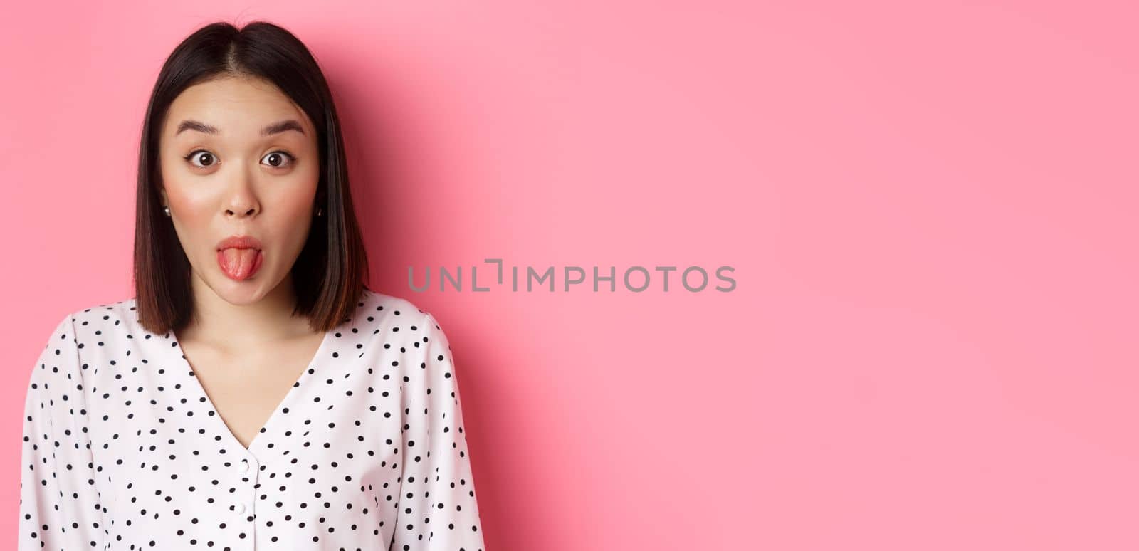 Beauty and lifestyle concept. Close-up of funny and cute asian woman showing tongue, staring at camera silly, standing over pink background.