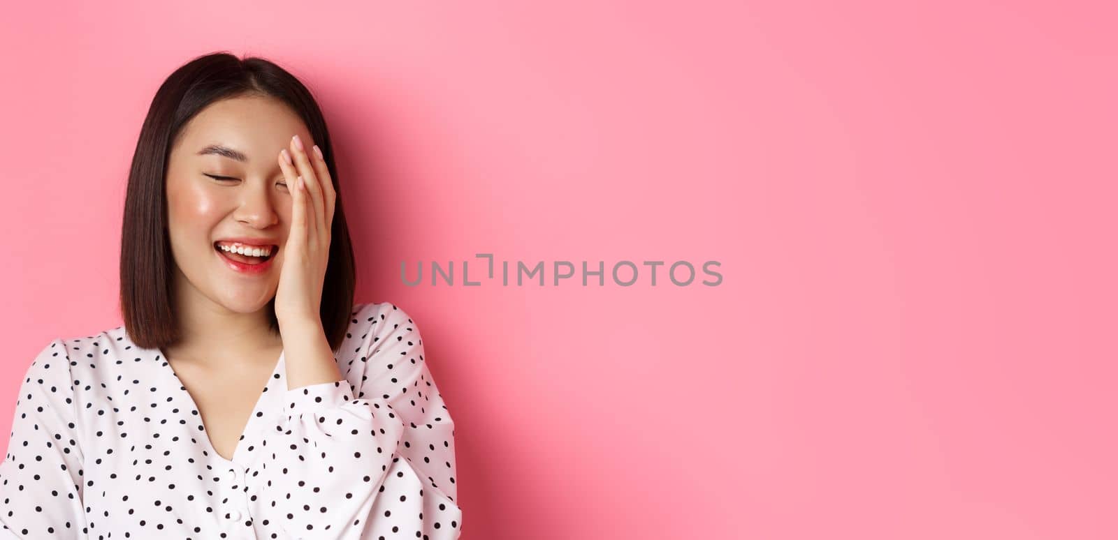 Beauty and lifestyle concept. Close-up of happy asian female laughing, looking happy and showing genuine emotions, standing over pink background.