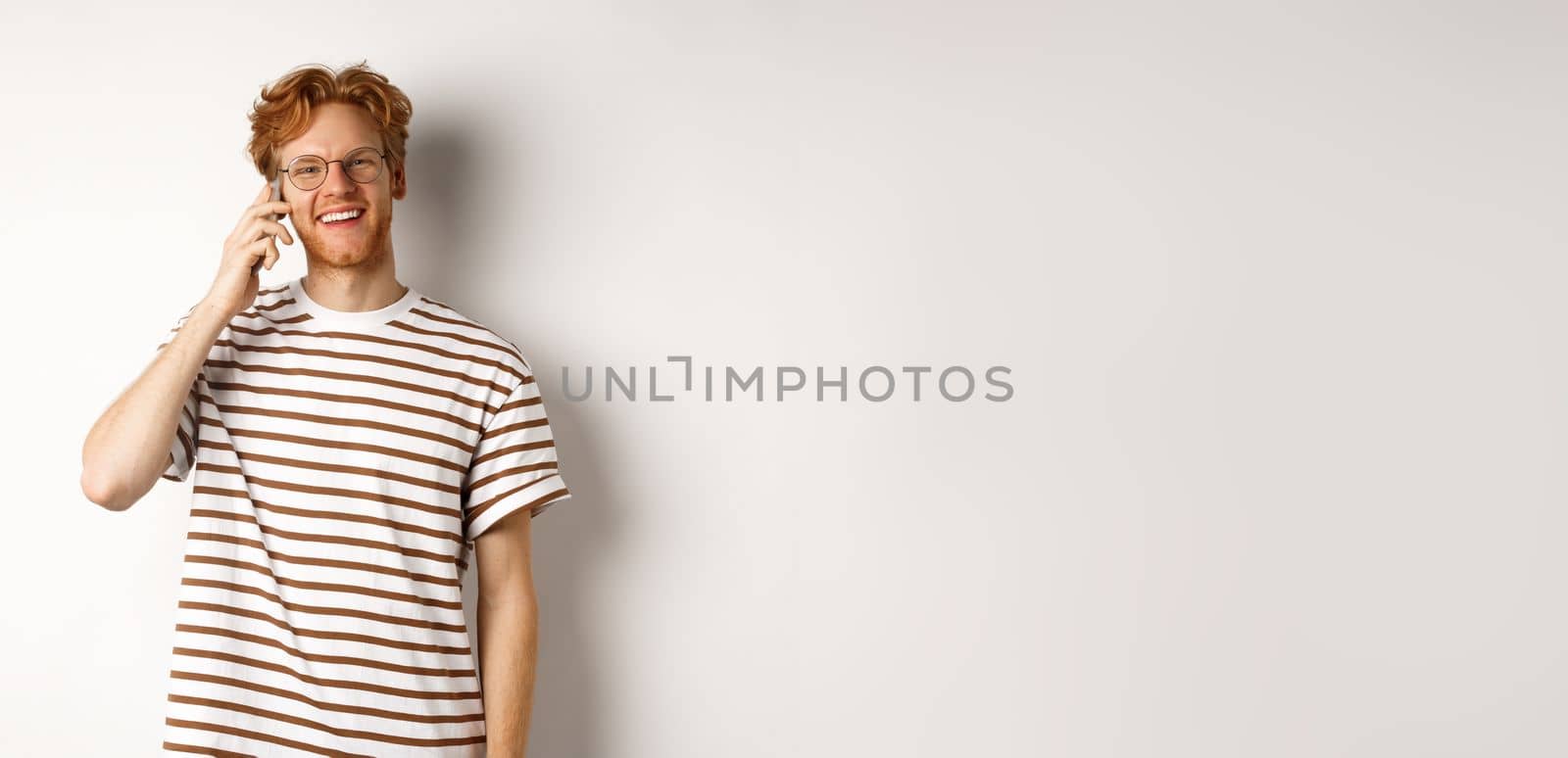 Hipster with red hair and glasses talking on mobile phone, smiling during conversation, white background.