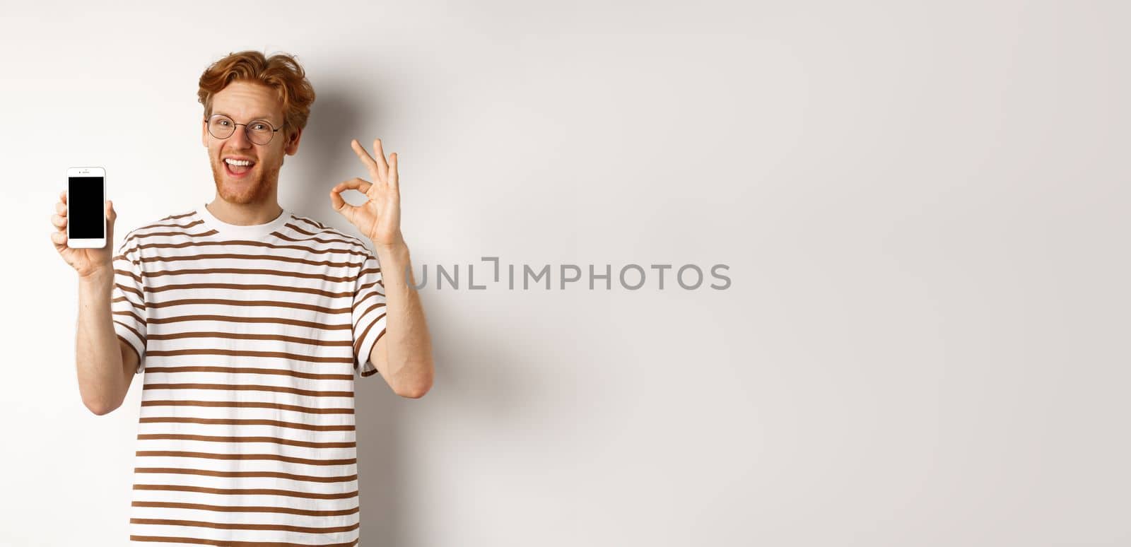 Technology and e-commerce concept. Young man with red hair showing okay sign and blank smartphone screen, praising awesome app, standing over white background by Benzoix