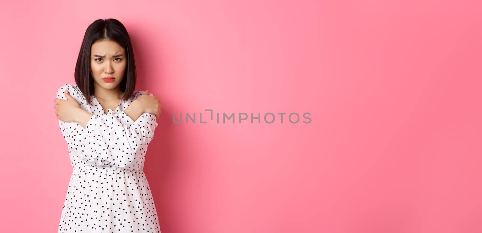 Timid and offended asian girl cross arms on chest, staring defensive and insulted at camera, standing in dress over pink background.