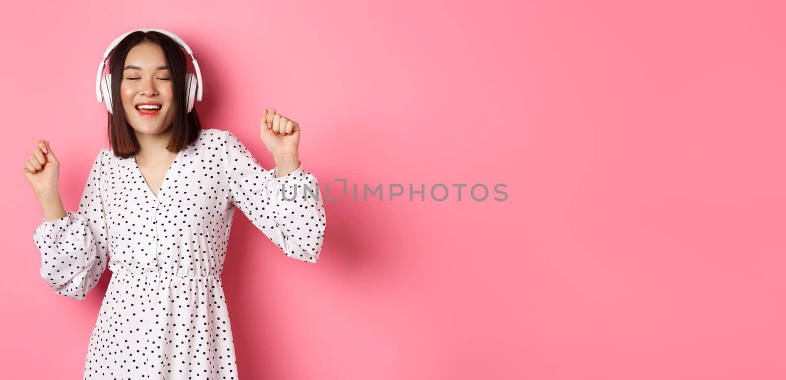 Happy young asian woman dancing and having fun, listening music in headphones, standing over pink background. Copy space