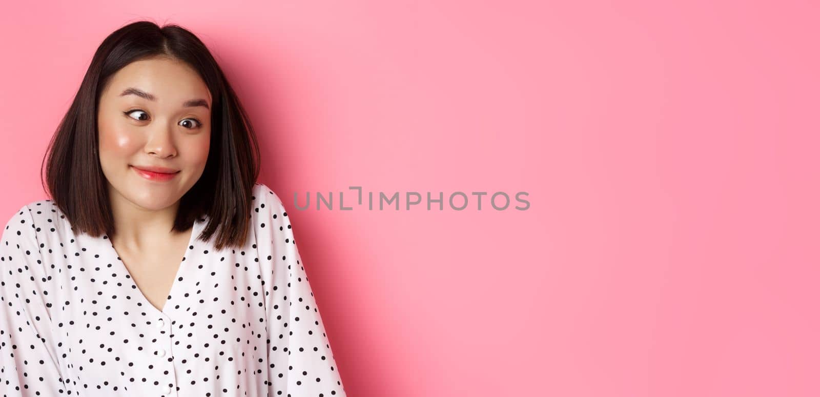 Beauty and lifestyle concept. Close-up of funny and cute asian woman squinting eyes and smiling, fool around at camera, standing over pink background.