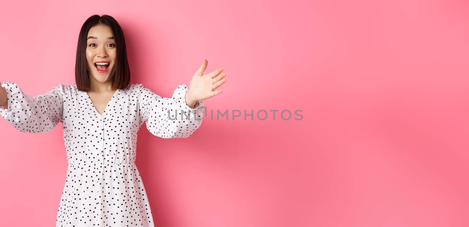 Happy asian girl stretching hands to camera, reaching for hug, smiling and wanting to hold something, standing over pink background in dress.