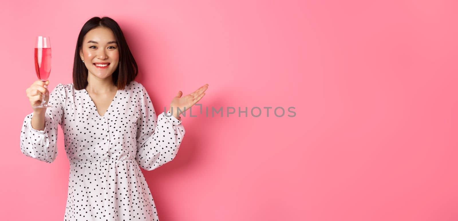 Beautiful asian woman raising glass of champagne and looking happy at camera, congratulating you on party, celebrating, standing over pink background.