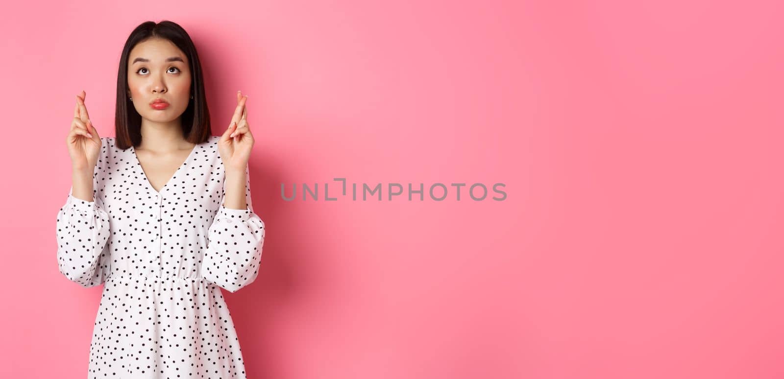 Cute asian woman making a wish, smiling happy and cross fingers for good luck, waiting over pink background.