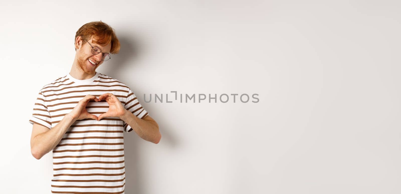 Valentines day. Happy boyfriend with red hair, smiling and showing heart gesture, I love you, standing over white background by Benzoix