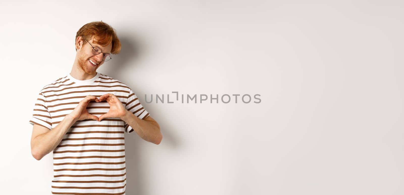 Valentines day. Happy boyfriend with red hair, smiling and showing heart gesture, I love you, standing over white background.