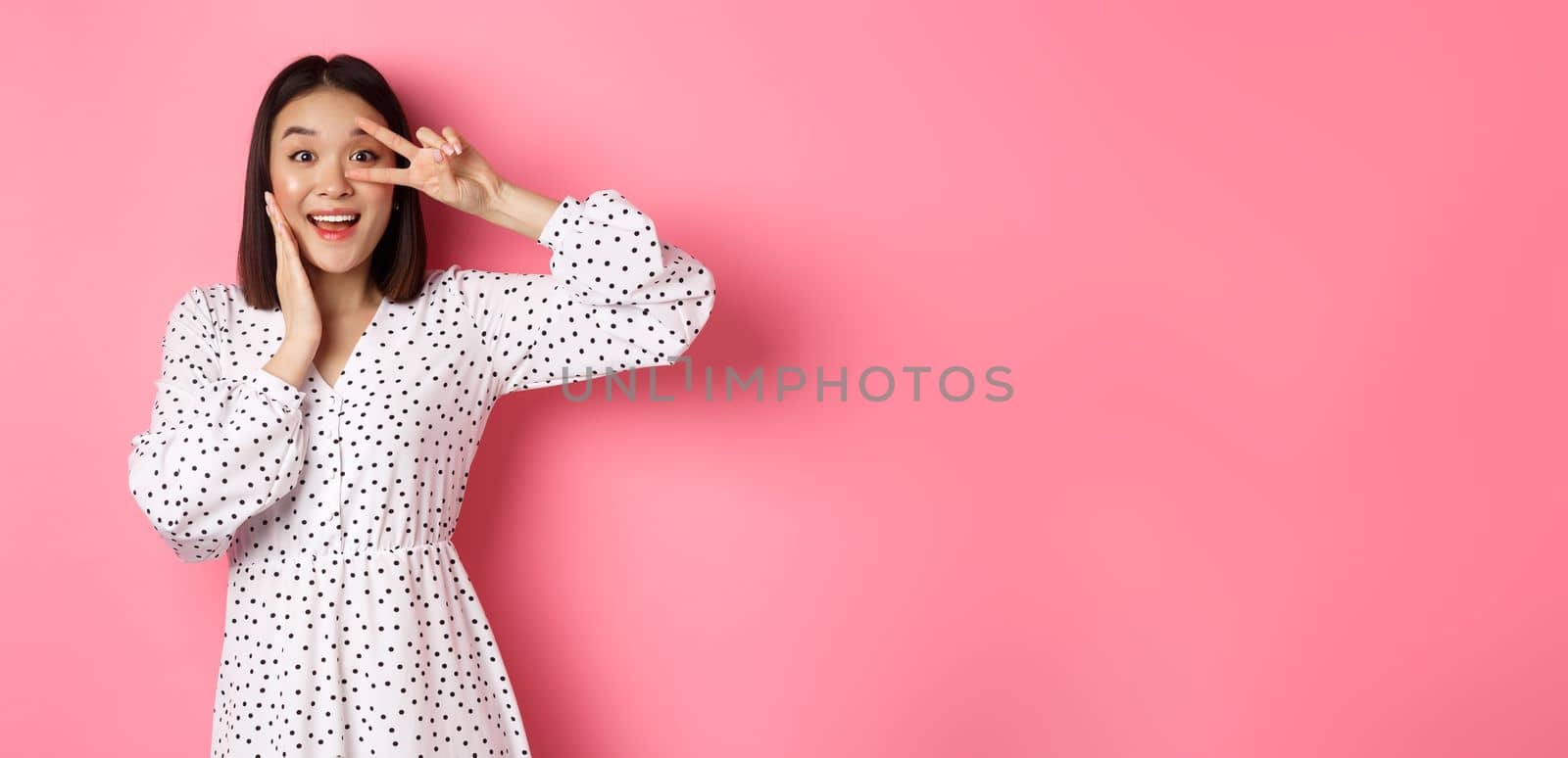 Beautiful and feminine asian woman showing kawaii sign on eye and looking amazed at camera, standing in dress over pink background.