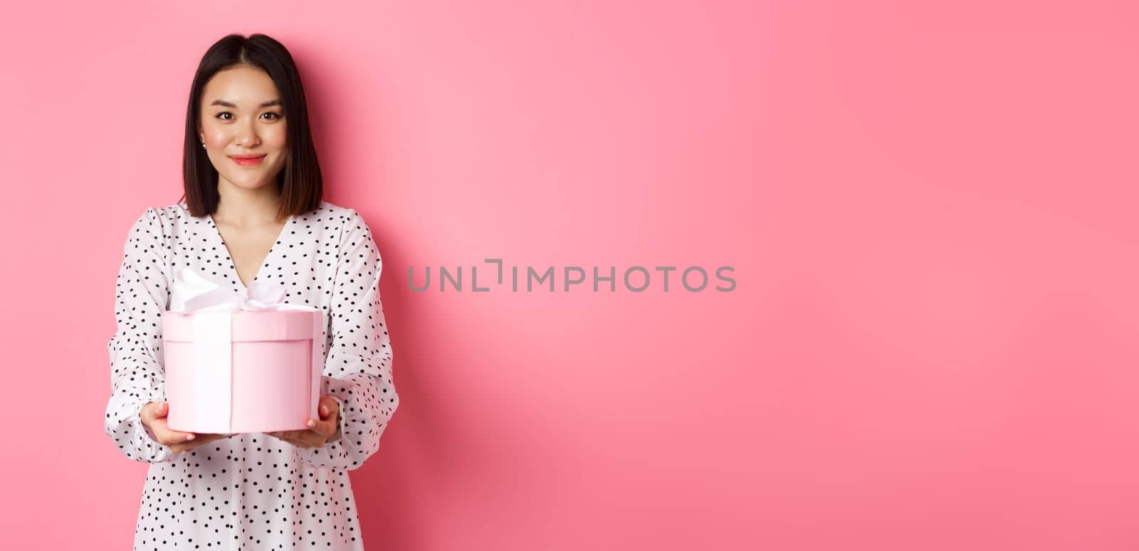 Beautiful asian woman wishing happy holidays, giving you gift in cute box, standing against pink background.