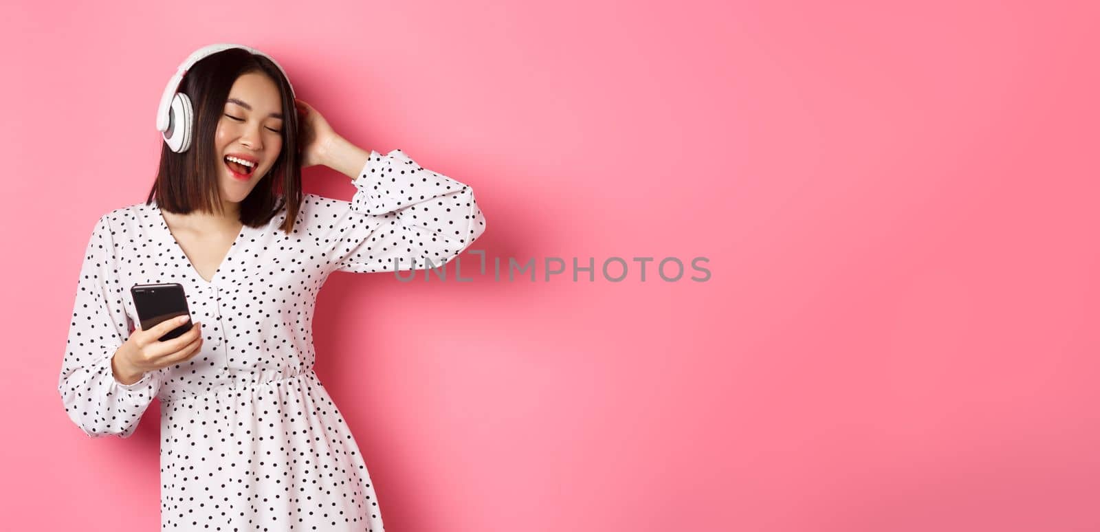 Carefree asian woman having fun, dancing and listening music in headphones, holding mobile phone, standing in dress over pink background by Benzoix