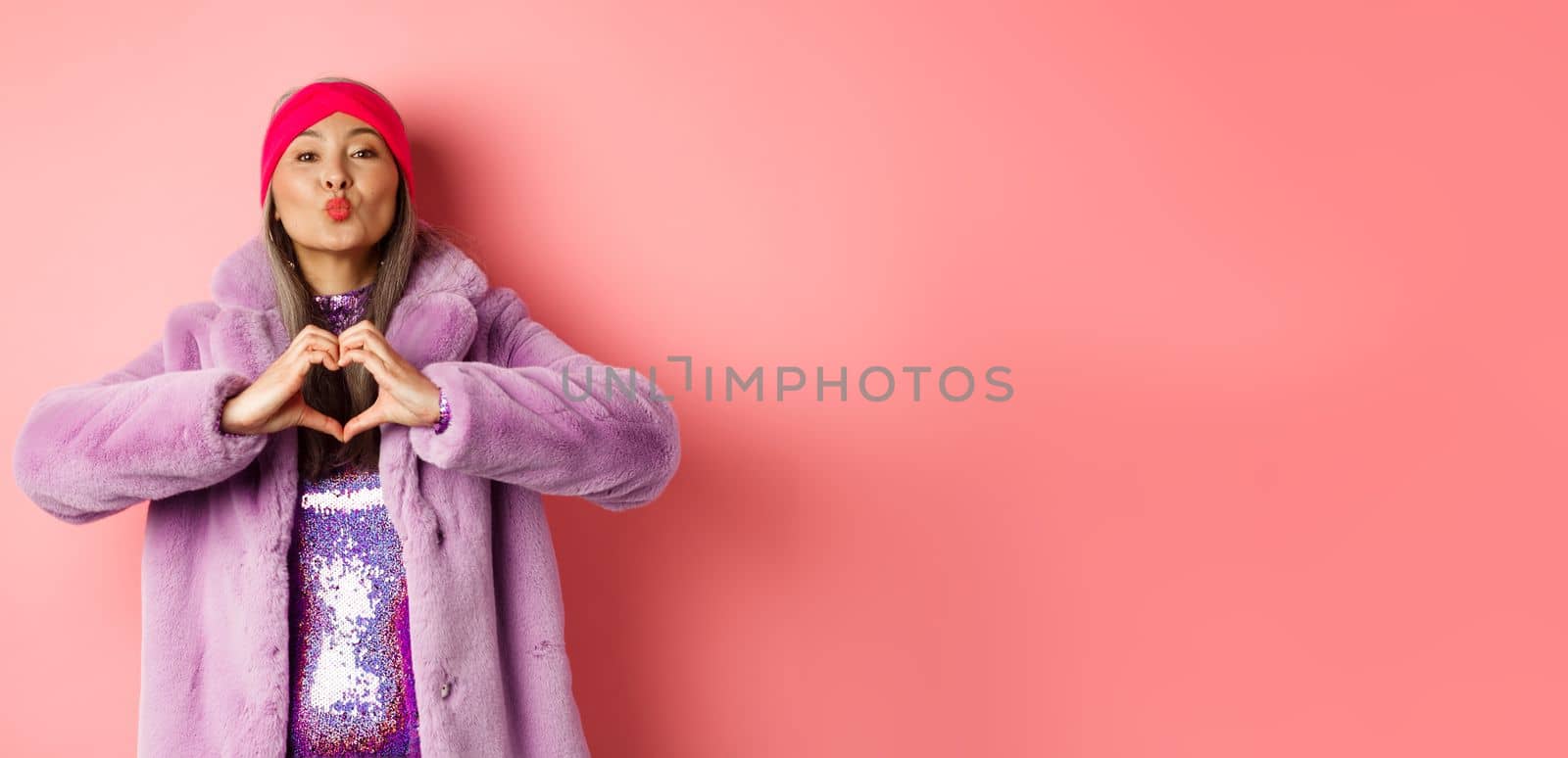 Romance and valentines day. Happy asian senior woman showing heart sign, I love you gesture, pucker lips for kiss, standing over pink background by Benzoix