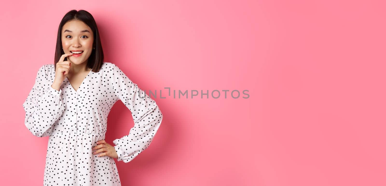 Image of beautiful young lady in dress, biting finger and smiling, staring with temptation and amazement, want buy something, standing over pink background.