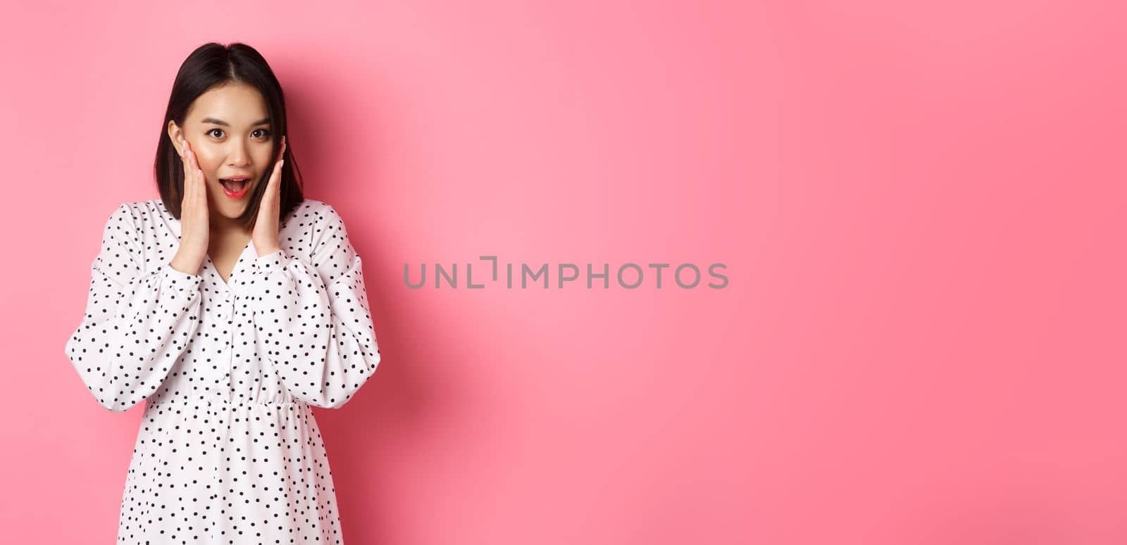 Young asian woman looking intrigued, gossiping and saying wow with curious face, looking interested at camera, standing over pink background.