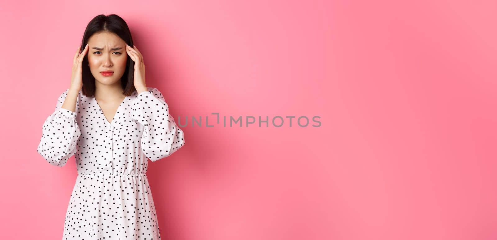 Image of upset asian woman having headache, feeling unwell or dizzy, close eyes and massaging head, suffering migraine, standing over pink background by Benzoix