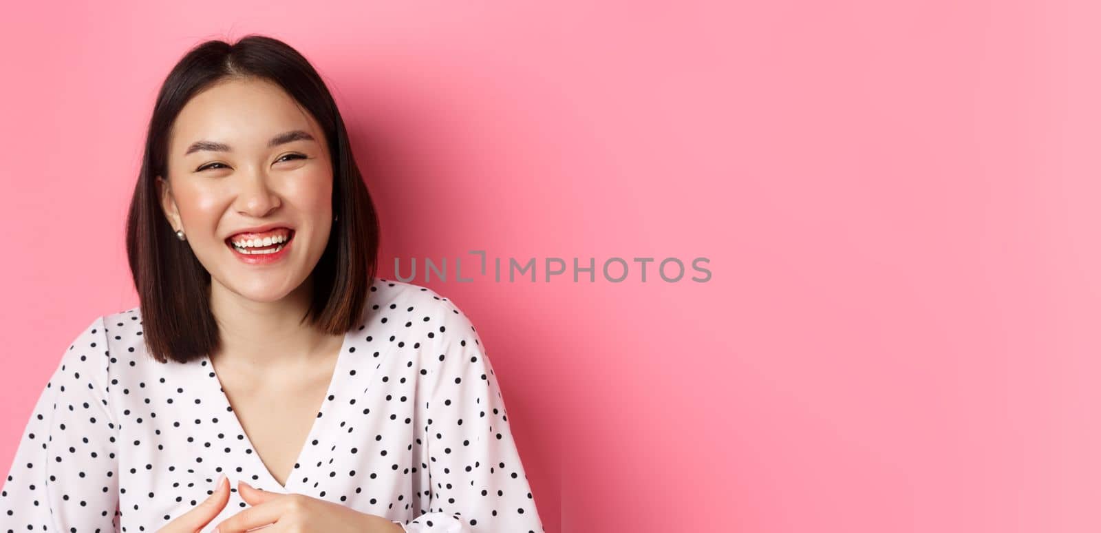 Beauty and lifestyle concept. Close-up of happy asian woman laughing and having fun, standing over pink background. Copy space
